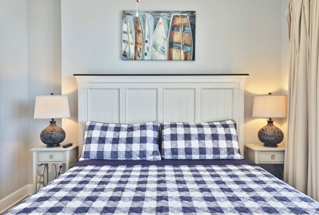 A neatly made bed with a checkered bedspread, flanked by two bedside tables with lamps, a white headboard, and a wall-mounted artwork above.