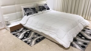 A neatly made bed with a white duvet, a headboard, and two patterned rugs on a tiled floor.
