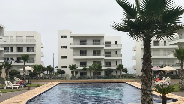 A swimming pool with lounge chairs and palm trees, surrounded by multi-story buildings.