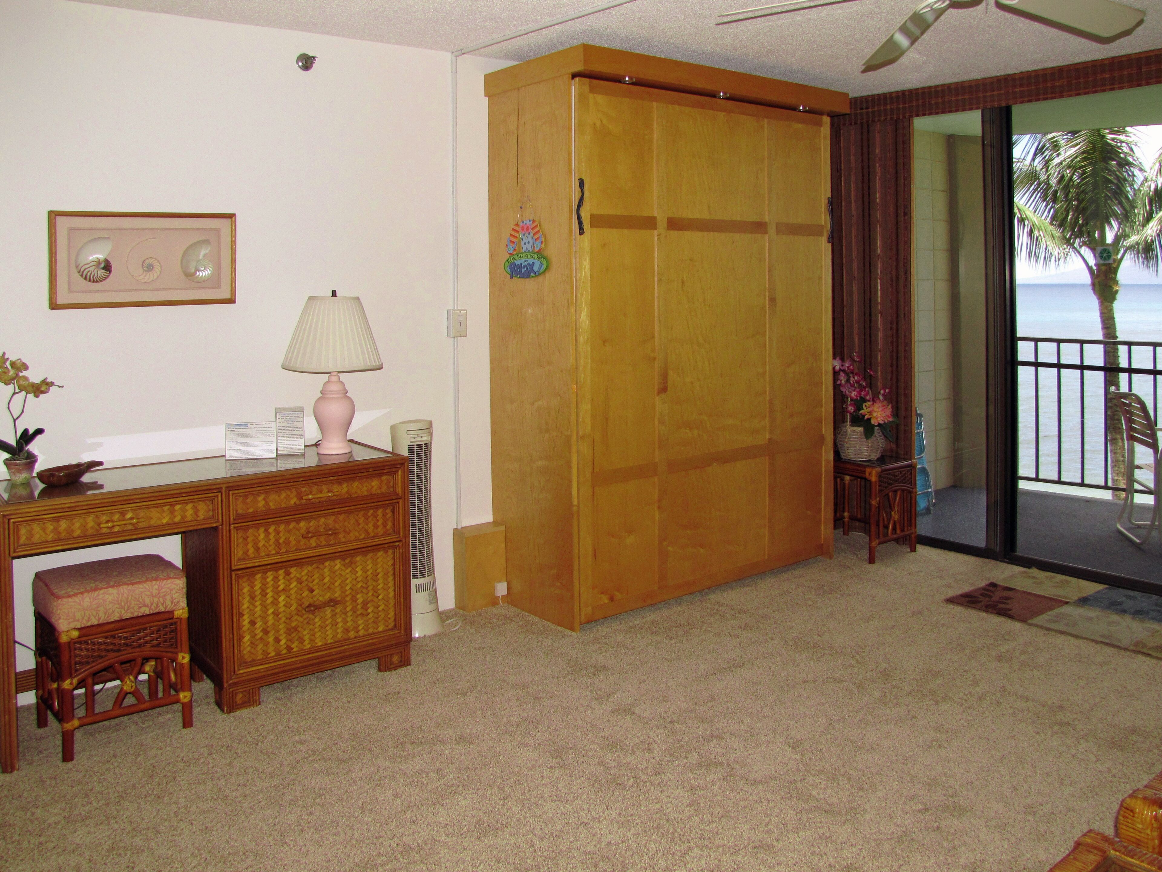 Une pièce avec une armoire en bois, un bureau avec une lampe, une chaise et une vue sur l’océan par une porte coulissante.
