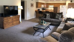 A living room with a TV, sofa, and coffee table. There is a kitchen in the background with wooden cabinets and a refrigerator.