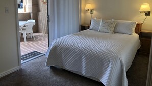 A bedroom with a large bed, two bedside lamps, and a view through a sliding door to an outdoor patio with a table and chairs.