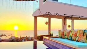 Una piscina infinita con vistas a la puesta de sol sobre el océano, un área de descanso con cojines de colores y un bar con botellas.