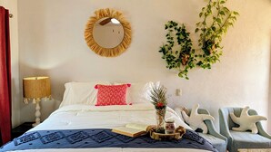 Un dormitorio ordenado con cama, lámpara de noche, silla y adornos en la pared, incluyendo un espejo y plantas.