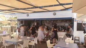 An outdoor dining area with tables and chairs under a canopy, a bar area with bottles and a chalkboard menu, and a view of the surrounding landscape. - Manilva Playa luxury apartment with sea view near the beach (Manilva)