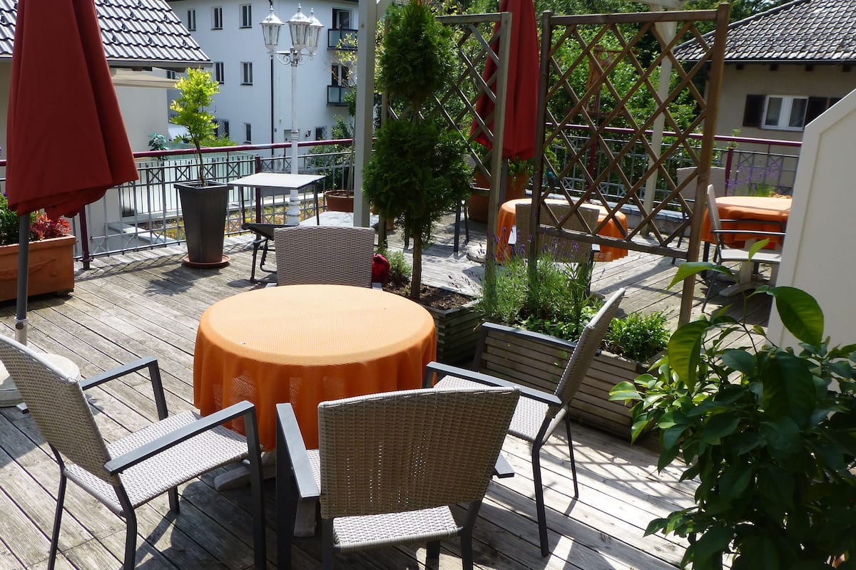 Una terraza de madera con mobiliario exterior, que incluye una mesa redonda y sillas, rodeada de macetas con plantas y un paraguas rojo.
