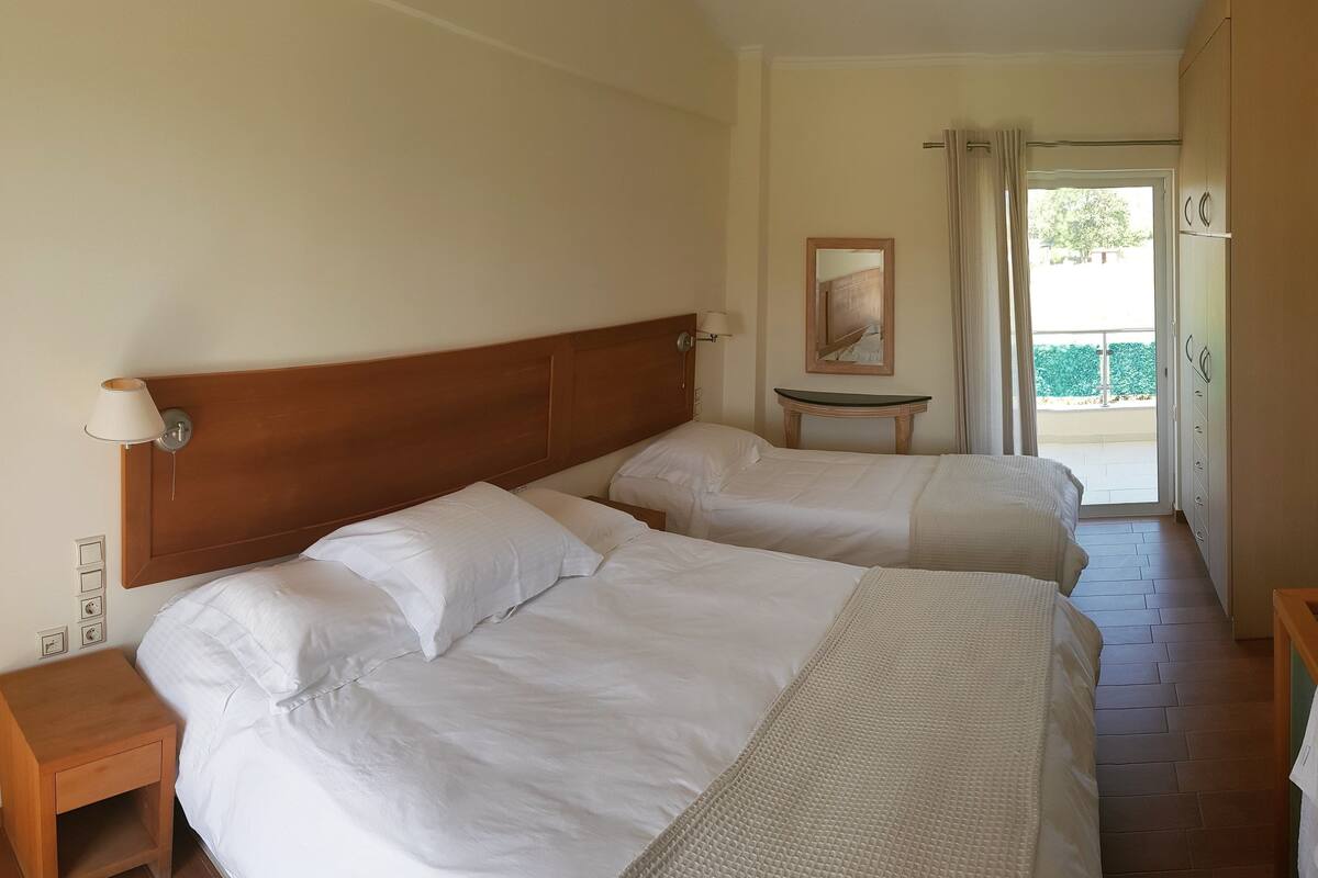 A hotel room with two beds, a small table, a TV, and a view of the outdoors.