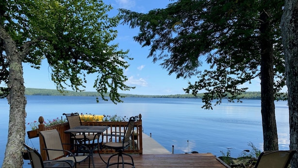 Eine Holzterrasse mit Gartenmöbeln, die auf einen ruhigen See mit umliegenden Bäumen blickt.
