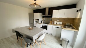 Een kleine keuken met witte kastjes, een magnetron, een fornuis en een vaatwasser. Er is een eettafel met twee stoelen en een hanglamp.