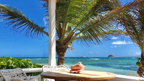 A wooden table with a seashell, white wicker chairs, and a view of the ocean with a distant island. - Two for One! Charming Cottage on hillside with private lounge on Turtle Beach! (BASSETERRE)