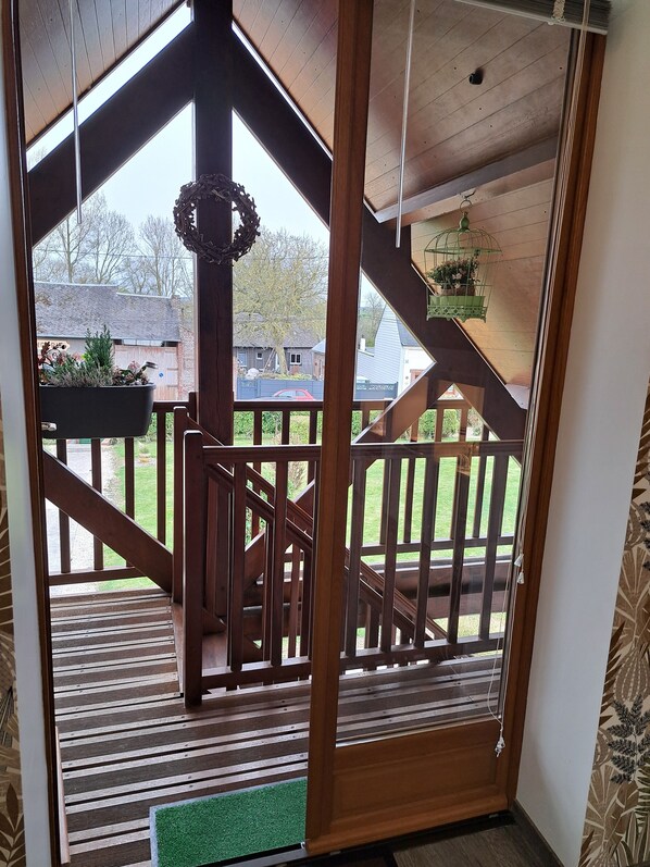 Una escalera de madera conduce a una terraza con barandilla, un farol colgante y vistas a un patio trasero con una casa y un coche.