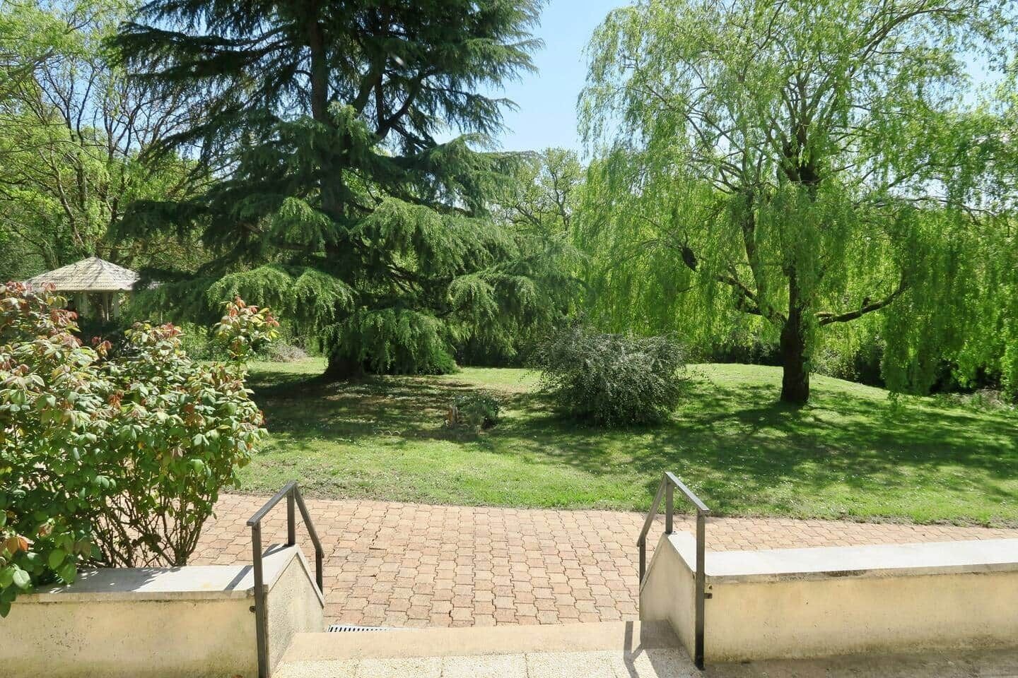 Un chemin pavé mène à un parc où se trouvent des arbres et un kiosque.