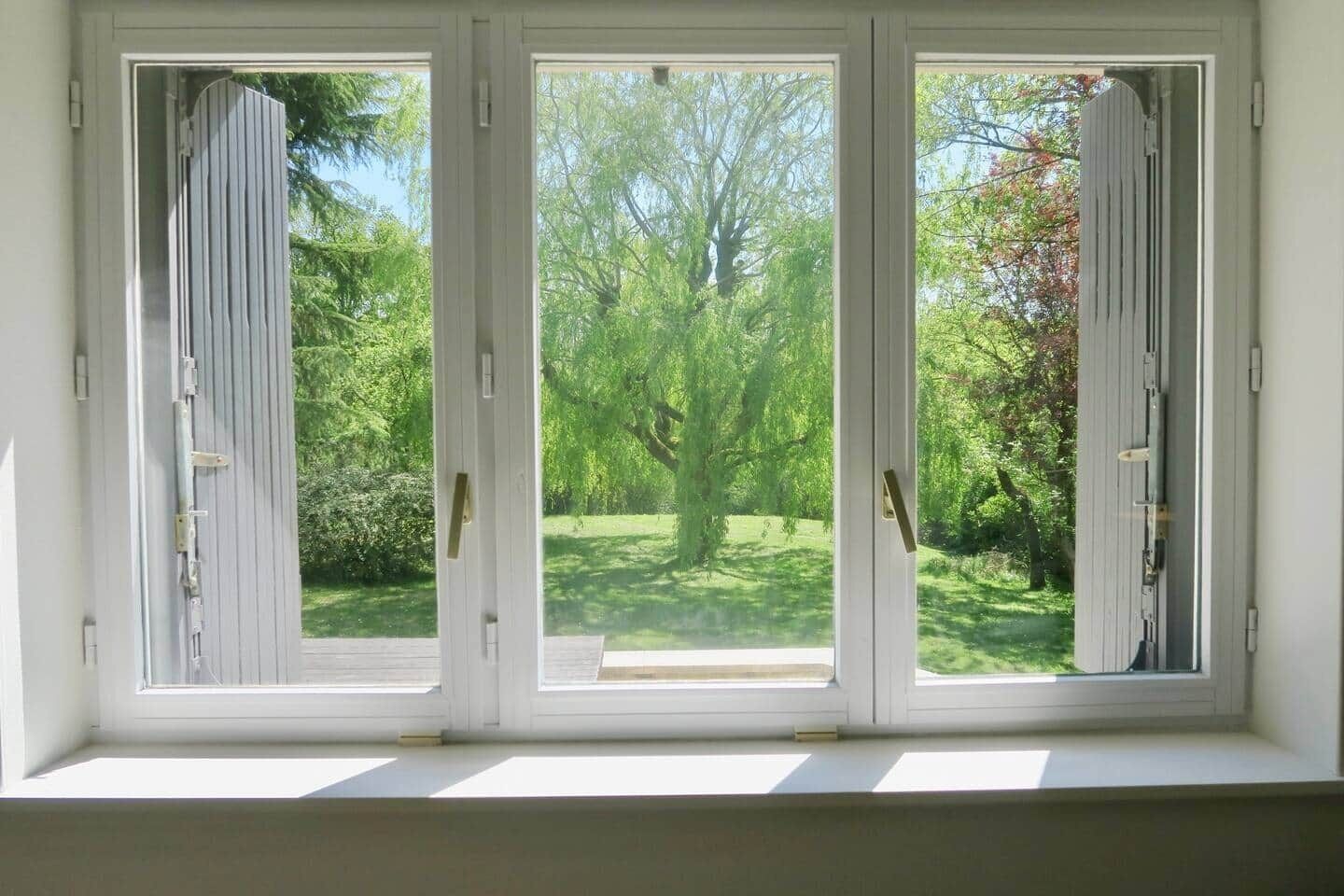 A window with three white-framed glass panes, each with a vertical metal handle, overlooking a green landscape with trees and a lawn.
