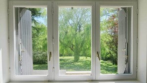 Una ventana con tres paneles de vidrio de marco blanco, cada uno con un tirador metálico vertical, que da a un paisaje verde con árboles y césped.