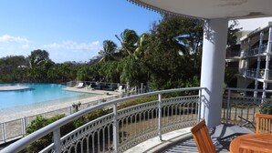 A balcony with a pool view, white railing, and a wooden table with a cushion.