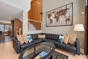 A modern living room with a curved glass coffee table, a sectional sofa with patterned cushions, and a large world map artwork on the wall. - Tremblant Prestige - Clairiere 406 (Mont-Tremblant)