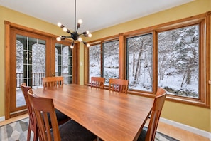 A dining room with a large wooden table and six chairs, large windows with a snowy forest view, and a modern chandelier. - Tremblant Prestige - Clairiere 406 (Mont-Tremblant)