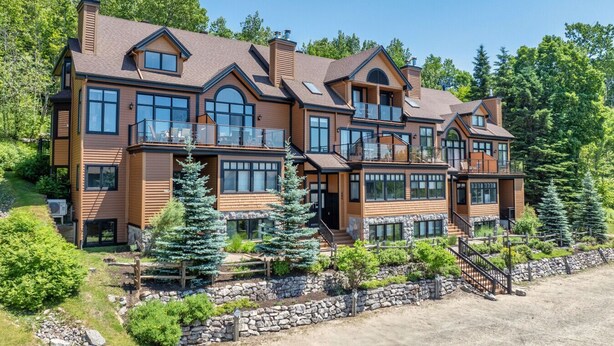 A multi-story wooden house with large windows and balconies, surrounded by greenery and a stone wall.