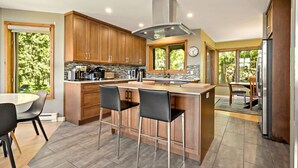 A modern kitchen with wooden cabinets, a central island with bar stools, a stainless steel range hood, and a large window providing a view of the outdoors.