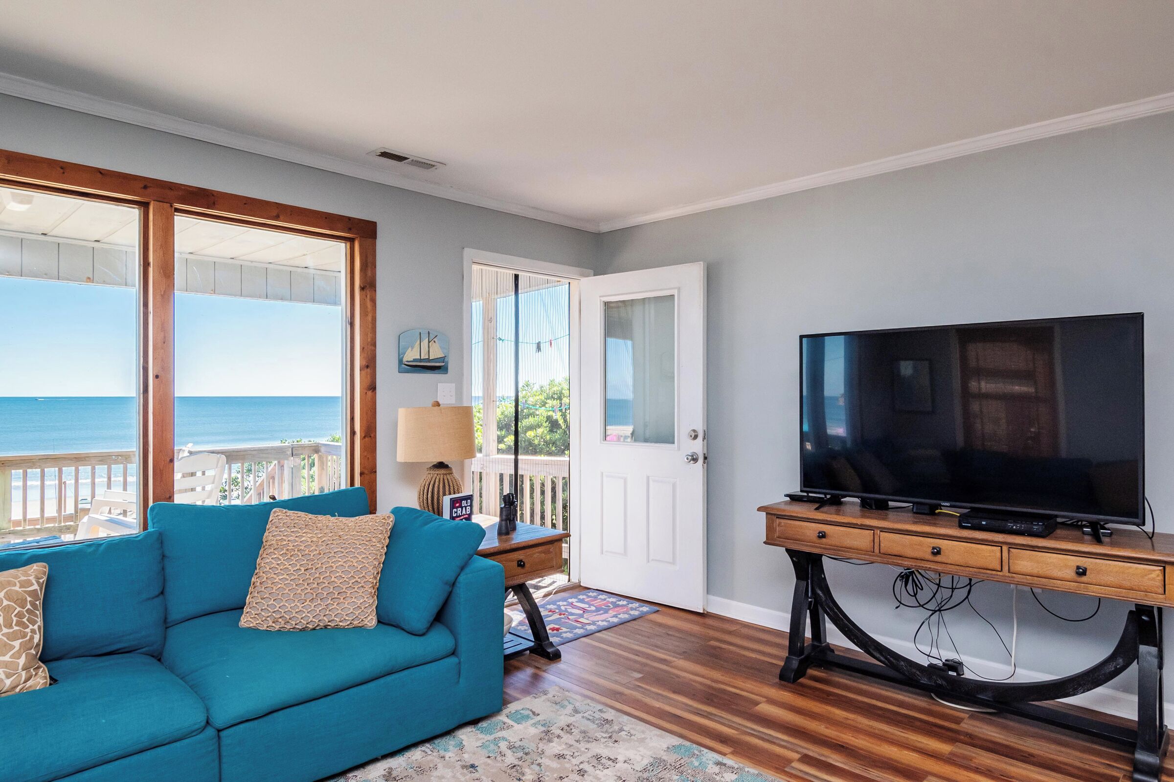 A living room with a blue sofa, a television on a wooden stand, and a view of the ocean through large windows.
