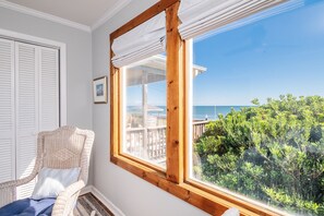 Quarto com uma grande janela com vista para um deck e para o oceano, uma cadeira de vime com almofada, e uma porta branca com persianas.