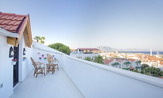 A balcony with wicker chairs and potted plants, overlooking a coastal town with red-roofed buildings and a clear sky.