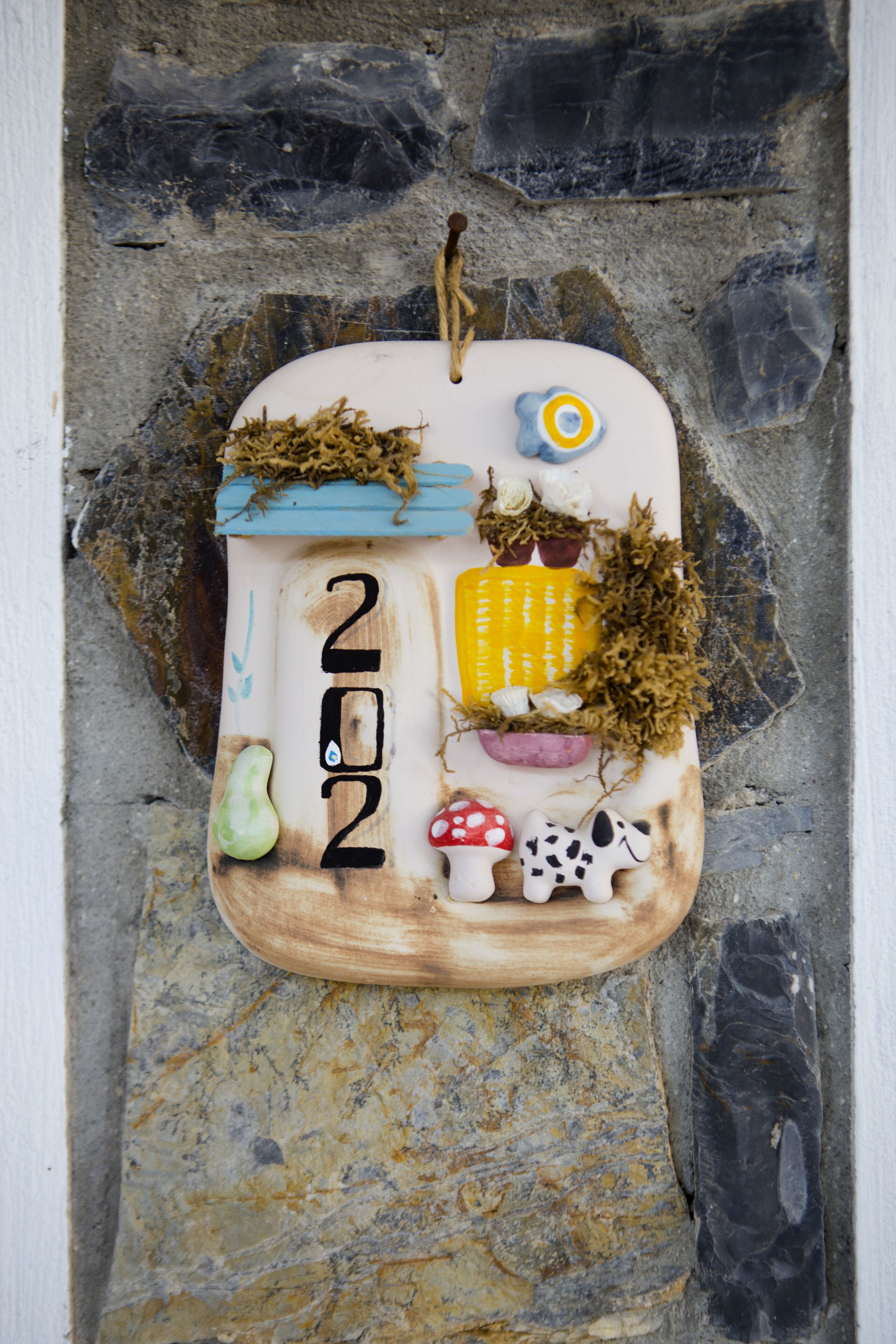 Een decoratief houten plaquette met een rustiek ontwerp, waarop het cijfer '202', een blauwe en gele bloem, een paddenstoel en een koe afgebeeld zijn.