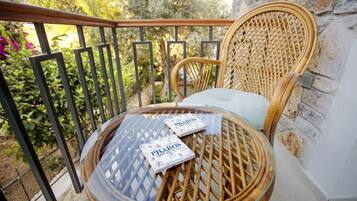 Une chaise en osier avec un coussin, sur un balcon avec une table en verre, deux cartes PHAROS et un mur en pierre.