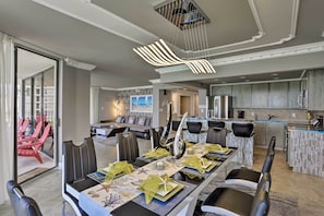 Un espace repas moderne avec une longue table dressée pour six personnes, une cuisine aux meubles gris et une vue sur un balcon agrémenté de chaises rouges.