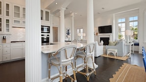 A spacious kitchen with white cabinetry, a central island with bar stools, a dining area with a sofa and chairs, and a fireplace. - Spectacular Ocean Views! Immaculate Family Oceanfront Home, Pool, Rooftop Deck! Emmeline Cottage (Saint Simons Island)