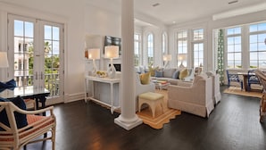 A spacious living room with a large window, a white sofa, a coffee table, and a dining area with a table and chairs. - Spectacular Ocean Views! Immaculate Family Oceanfront Home, Pool, Rooftop Deck! Emmeline Cottage (Saint Simons Island)