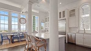 A spacious kitchen with white cabinetry, a central island, and a dining area with blue chairs. - Spectacular Ocean Views! Immaculate Family Oceanfront Home, Pool, Rooftop Deck! Emmeline Cottage (Saint Simons Island)