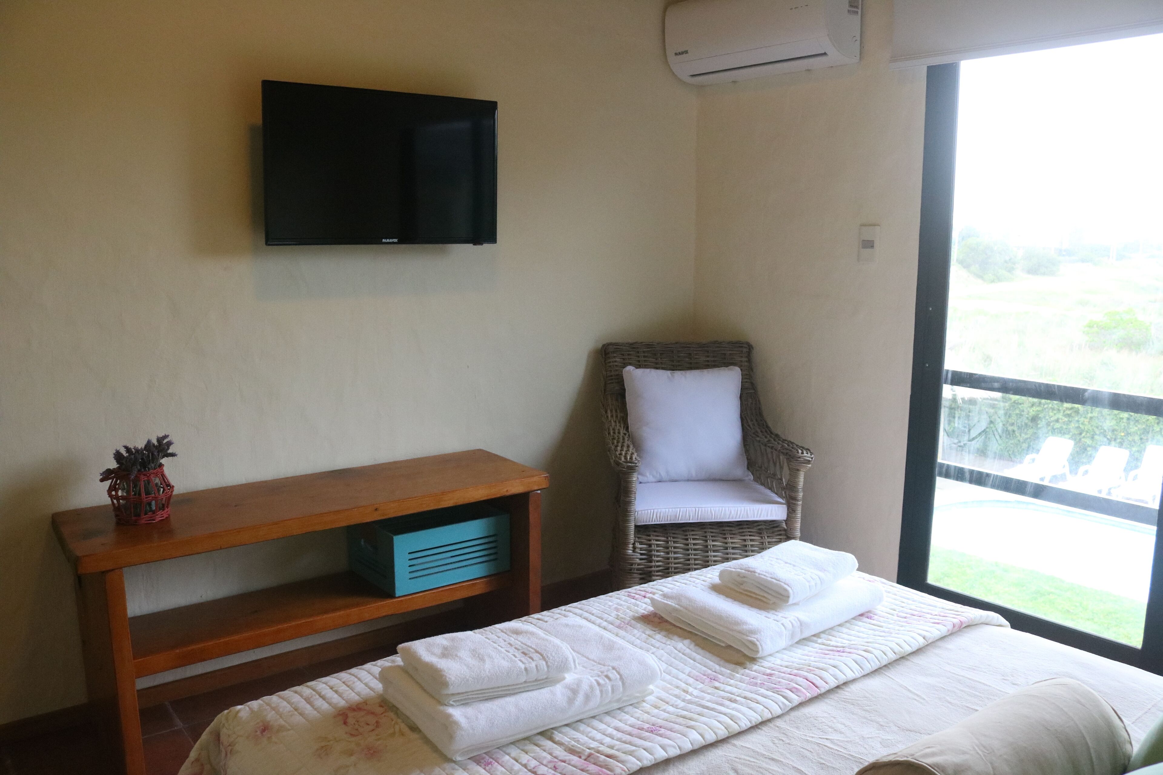 Un dormitorio con cama, televisor en la pared, una silla de mimbre y ventana con vistas.