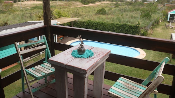 A wooden deck with a small table and chairs, overlooking a swimming pool and a grassy area. - Widow's Inn - Superior Double (Punta del Diablo, Rocha)