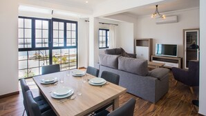 Ein modernes Wohnzimmer mit Essbereich, großen Fenstern, einem grauen Sofa und einem Flachbildfernseher.