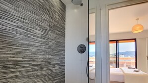 A modern bathroom with a textured wall, a glass shower enclosure, and a view of a bedroom with a bed and a towel.