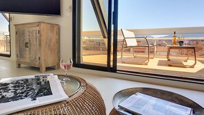 A room with a large window, a wooden cabinet, a wicker chair, a glass table with a wine glass, and a newspaper.