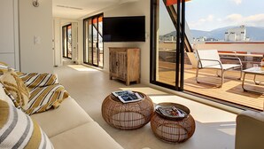 A modern living room with a sofa, two wicker side tables, a wooden cabinet, and a TV. Large windows offer a view of the outdoors.