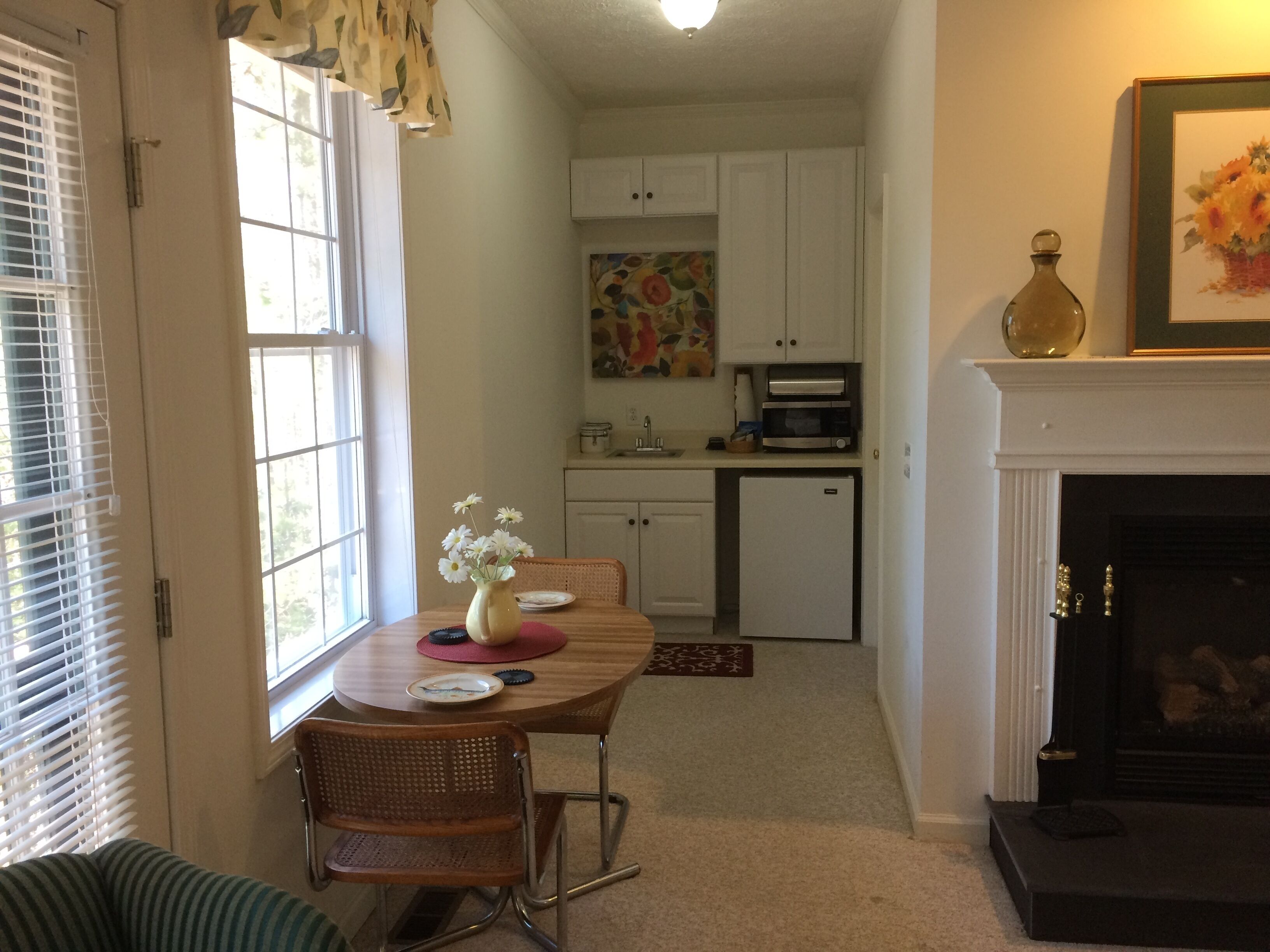 A dining area with a table, chairs, and a vase of flowers. There is a kitchen in the background with cabinets and appliances.