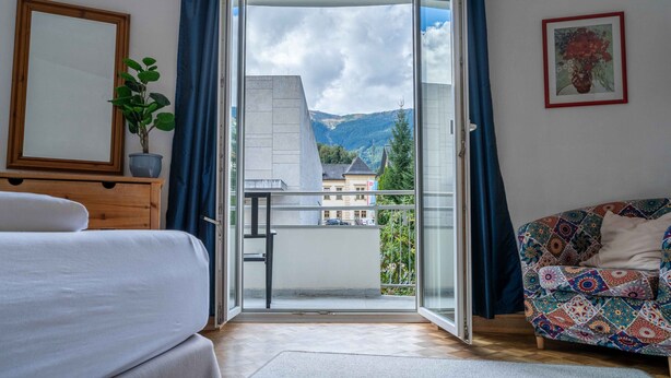 A bedroom with a bed, a dresser, a potted plant, a balcony with a view of mountains and buildings, and a colorful chair. - Living Eden - TOP 3, city center & balcony (Zell am See)