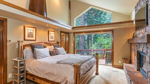 A spacious bedroom with a large bed, a stone fireplace, and a balcony with a view of trees. - 4BR Incline Village Home | Hot Tub, Game Room (Incline Village)