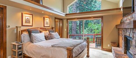A spacious bedroom with a large bed, a stone fireplace, and a balcony with a view of trees.