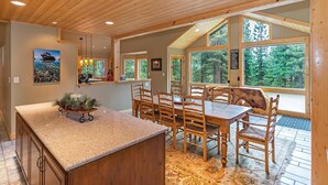 A spacious dining area with a large wooden table and chairs, a kitchen with a granite countertop, and large windows offering a view of the surrounding forest. - 4BR Incline Village Home | Hot Tub, Game Room (Incline Village)