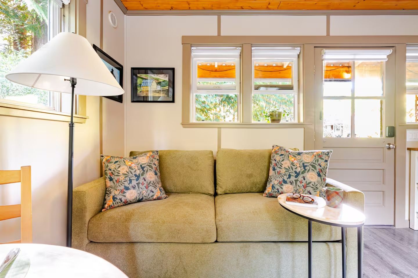 A room with a sofa, two pillows, a table with a cup and glasses, a lamp, and a window with a view of greenery.