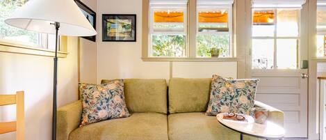 A room with a sofa, two pillows, a table with a cup and glasses, a lamp, and a window with a view of greenery.