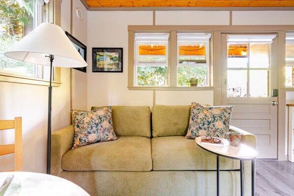 A room with a sofa, two pillows, a table with a cup and glasses, a lamp, and a window with a view of greenery.