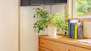Une pièce avec un plafond en bois, un téléviseur à écran plat fixé au mur, une commode en bois avec des tiroirs, un tabouret en osier et une fenêtre donnant sur un paysage verdoyant.