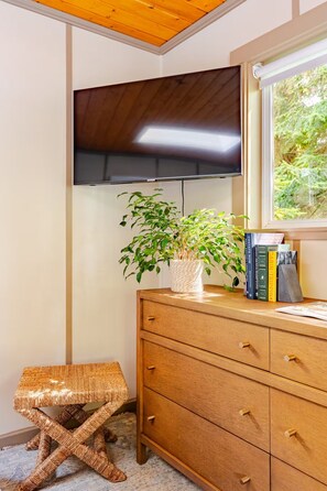 A room with a wooden ceiling, a flat-screen TV mounted on the wall, a wooden dresser with drawers, a woven stool, and a window with a view of greenery.