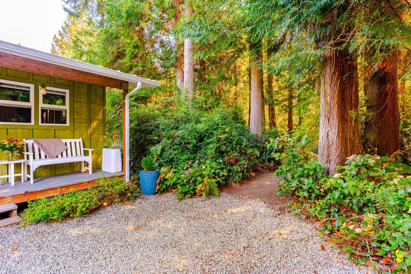 A cabin with a porch, a bench, and a gravel path leading into a forest.