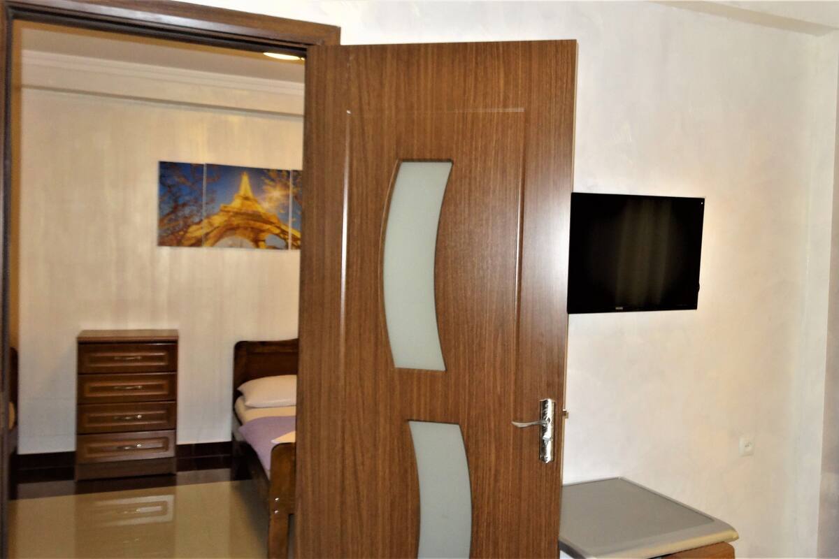 A hotel room with a wooden door, a bed, a desk, a chair, a television, and a painting on the wall.